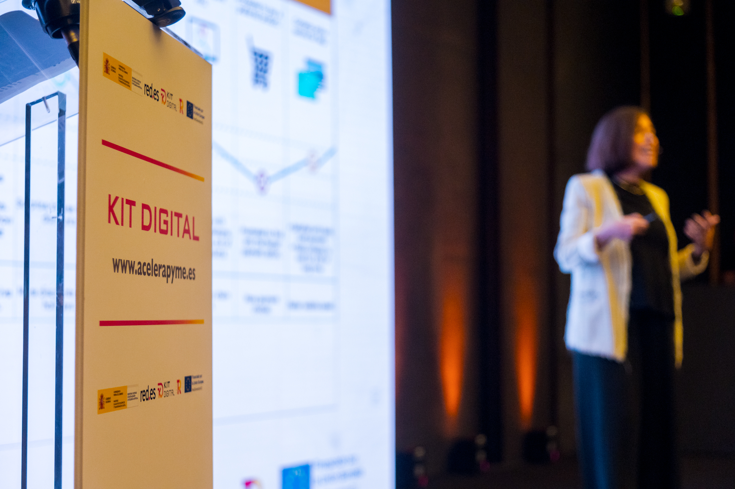 Atril con el texto ‘Kit Digital’ y la web www.acelerapyme.es en primer plano, junto a logotipos institucionales. Al fondo, una persona realiza una presentación frente a una pantalla con gráficos y esquemas iluminados.