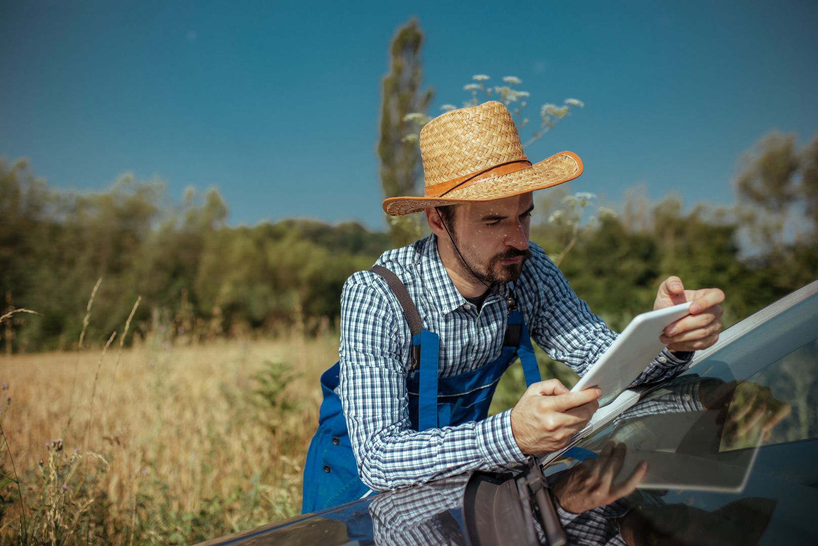 Persona con sombrero de paja y camisa a cuadros apoyada sobre un vehículo, utilizando una tableta en un entorno rural con vegetación y árboles al fondo bajo cielo despejado.