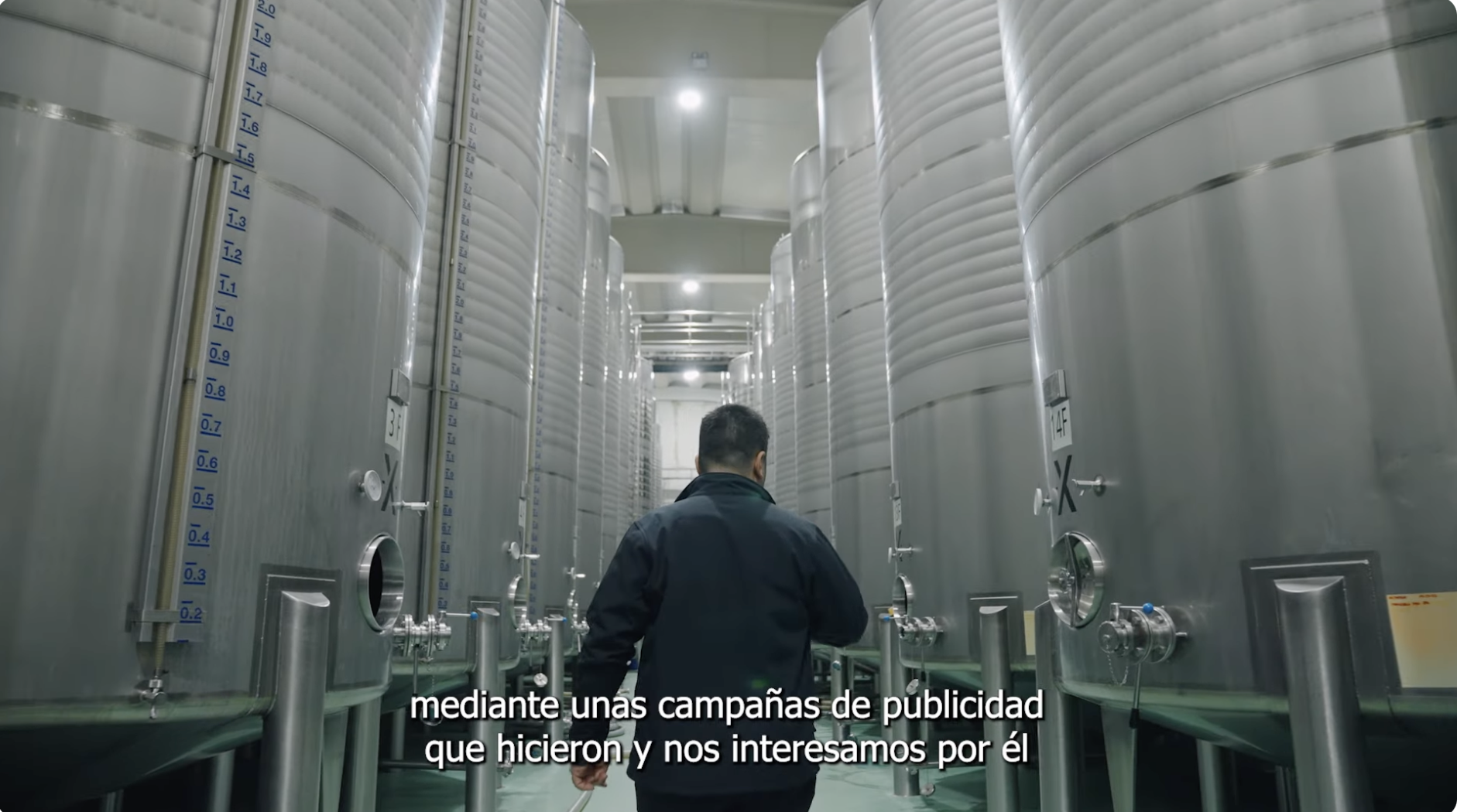 Persona caminando entre grandes depósitos metálicos en una planta industrial, con indicadores de nivel visibles en los tanques. En la parte inferior aparece el texto ‘mediante unas campañas de publicidad que hicieron y nos interesamos por él’.