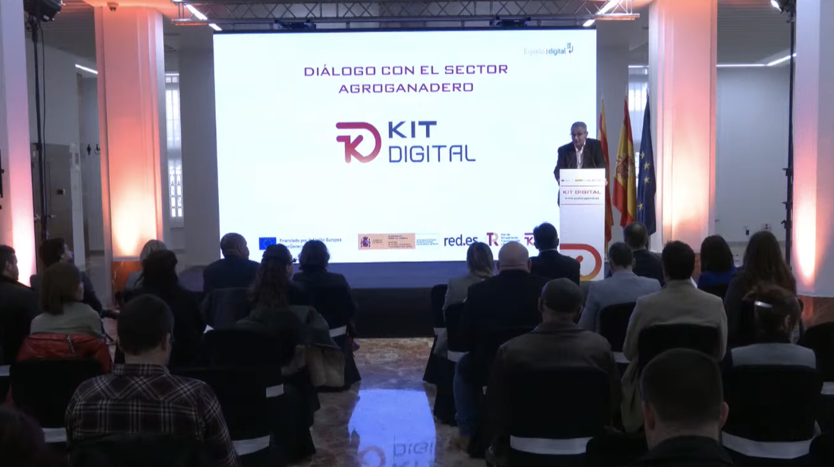 Evento del programa Kit Digital con público sentado frente a un escenario donde se muestra la pantalla con el texto ‘Diálogo con el sector agroganadero’ y el logotipo de Kit Digital, junto a banderas oficiales y un atril.