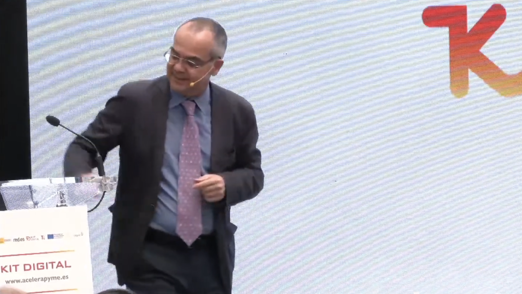 Persona de pie junto a un atril con micrófono durante una presentación sobre el programa Kit Digital, con el logotipo y la web ‘www.acelerapyme.es’ visibles en el atril y parte del logotipo en la pantalla de fondo.