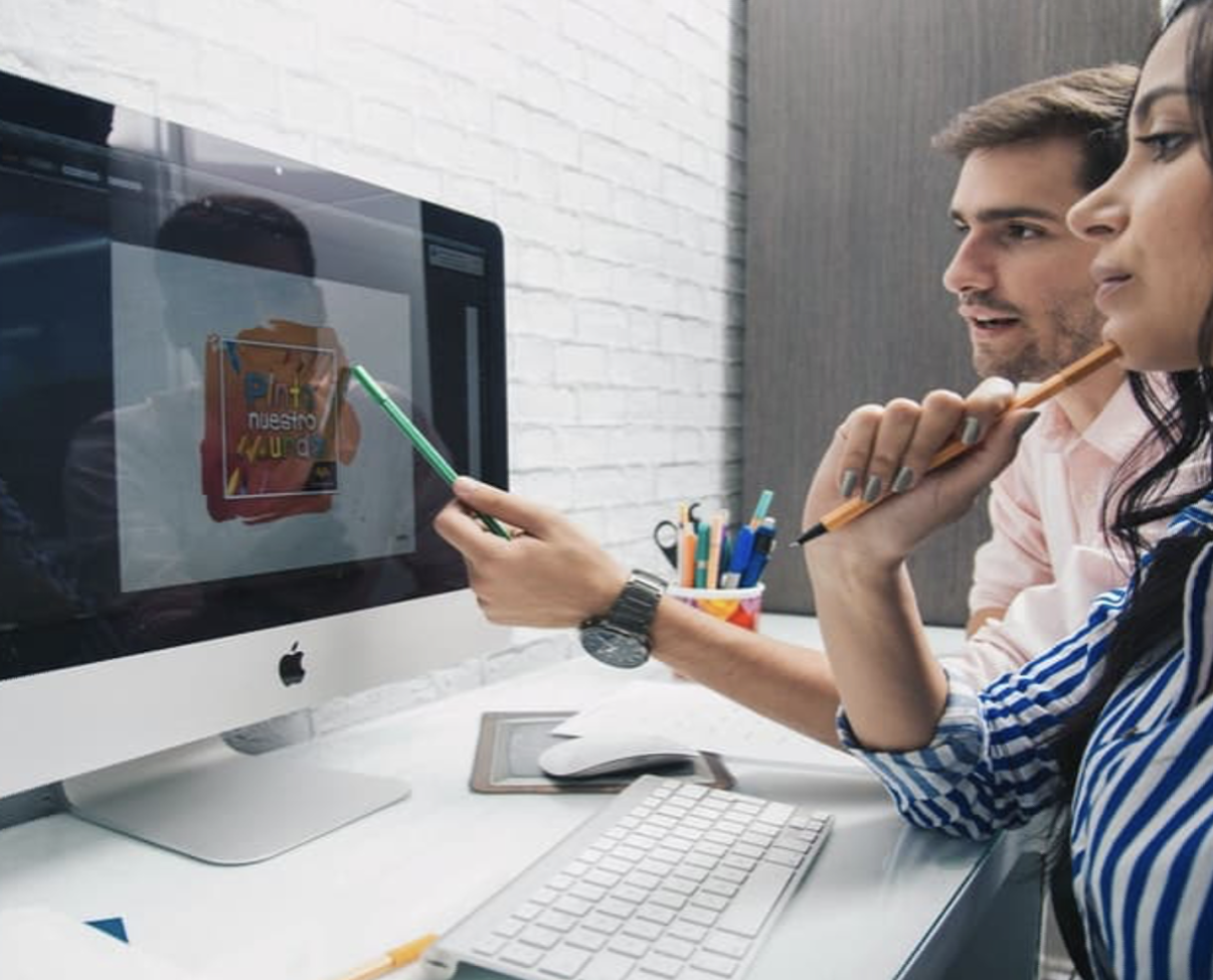 Dos personas colaboran en el diseño gráfico mostrado en la pantalla de un ordenador iMac, señalando elementos con lápices y un bolígrafo sobre un escritorio con teclado y accesorios.